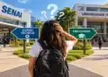 Jovem de mochila observa placas “Curso Técnico” e “Faculdade” diante do SENAI e universidade federal, simbolizando a dúvida entre cursos técnicos e faculdade.