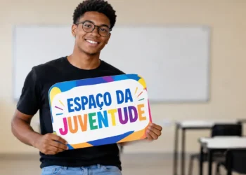 Jovem negro de óculos redondos e roupa preta segura placa colorida com o nome Espaço da Juventude em sala de aula.