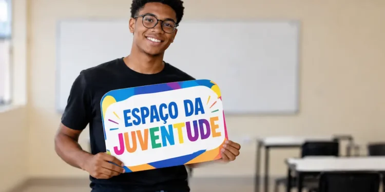 Jovem negro de óculos redondos e roupa preta segura placa colorida com o nome Espaço da Juventude em sala de aula.