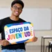 Jovem negro de óculos redondos e roupa preta segura placa colorida com o nome Espaço da Juventude em sala de aula.