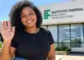 Mulher negra de blusa preta, calça jeans e acessórios azul-escuros sorri e acena para a câmera em frente ao prédio do IFMG Campus Ouro Preto, sob a luz do sol.