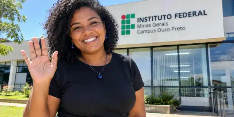 Mulher negra de blusa preta, calça jeans e acessórios azul-escuros sorri e acena para a câmera em frente ao prédio do IFMG Campus Ouro Preto, sob a luz do sol.