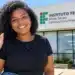 Mulher negra de blusa preta, calça jeans e acessórios azul-escuros sorri e acena para a câmera em frente ao prédio do IFMG Campus Ouro Preto, sob a luz do sol.