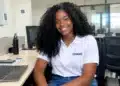 Jovem sorridente com cabelo preto, camiseta branca e calça jeans em ambiente de escritório.