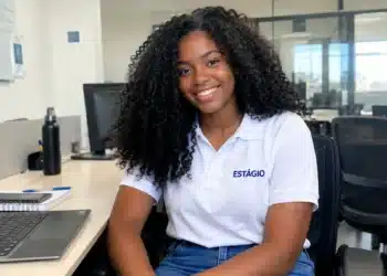 Jovem sorridente com cabelo preto, camiseta branca e calça jeans em ambiente de escritório.
