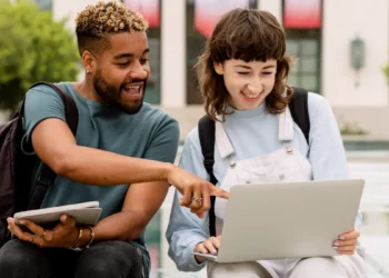 Jovens interagindo animados com notebook em área externa universitária.