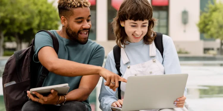Jovens interagindo animados com notebook em área externa universitária.