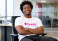 Homem negro de cabelo crespo, usando camiseta branca com o logo da Livelo e calça preta, sentado em uma cadeira de escritório, sorrindo de braços cruzados em um ambiente corporativo moderno e iluminado.