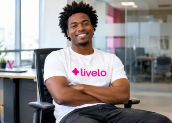 Homem negro de cabelo crespo, usando camiseta branca com o logo da Livelo e calça preta, sentado em uma cadeira de escritório, sorrindo de braços cruzados em um ambiente corporativo moderno e iluminado.