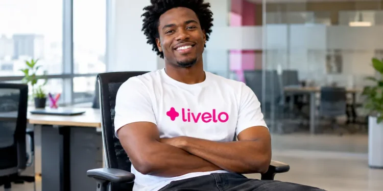 Homem negro de cabelo crespo, usando camiseta branca com o logo da Livelo e calça preta, sentado em uma cadeira de escritório, sorrindo de braços cruzados em um ambiente corporativo moderno e iluminado.