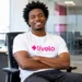 Homem negro de cabelo crespo, usando camiseta branca com o logo da Livelo e calça preta, sentado em uma cadeira de escritório, sorrindo de braços cruzados em um ambiente corporativo moderno e iluminado.