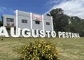 Letreiro de Augusto Pestana em área gramada, com flores amarelas e árvores ao fundo.