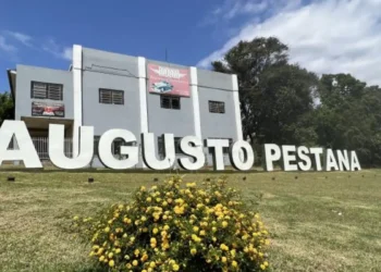 Letreiro de Augusto Pestana em área gramada, com flores amarelas e árvores ao fundo.