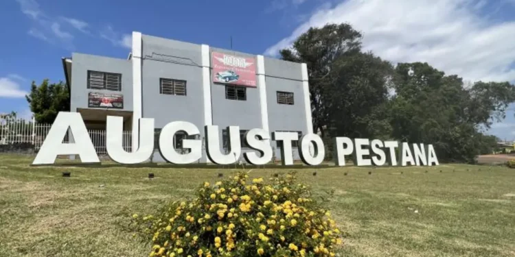 Letreiro de Augusto Pestana em área gramada, com flores amarelas e árvores ao fundo.
