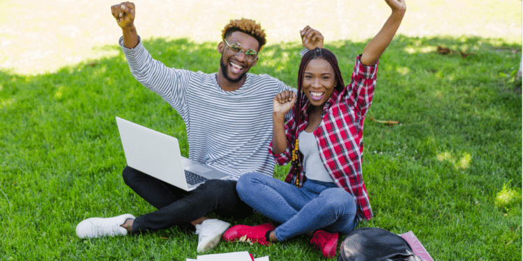 Dois jovens sentados na grama, sorrindo com braços erguidos, diante de notebook aberto.