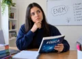 Estudante pensativa com livro de Física em quarto de estudos.