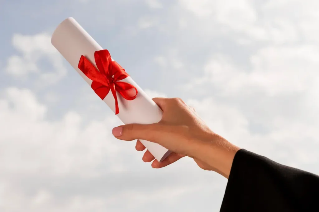 Mão feminina segurando diploma enrolado com laço vermelho contra o céu.