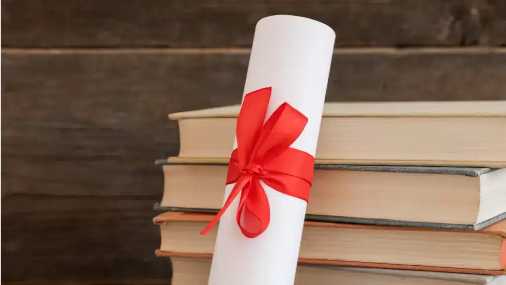 Diploma enrolado com fita vermelha ao lado de livros empilhados.
