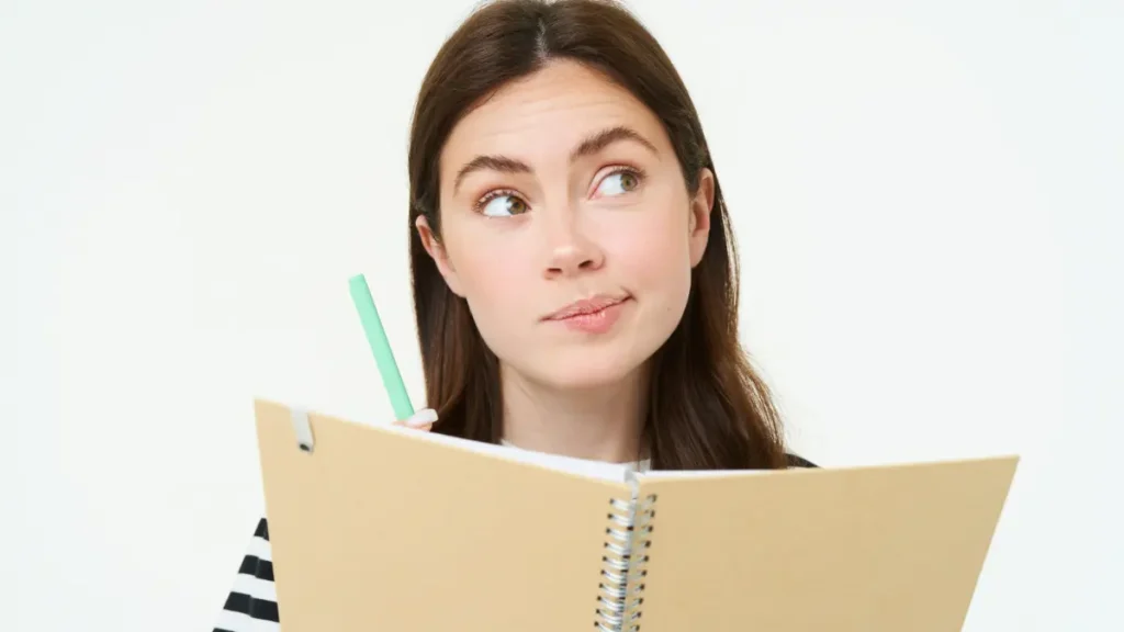 Jovem pensativa segurando caderno e caneta em fundo branco.