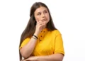 Jovem pensativa com camiseta amarela em fundo branco.
