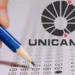 Mão segurando lápis azul preenche cartão-resposta ao lado do logotipo da Unicamp.