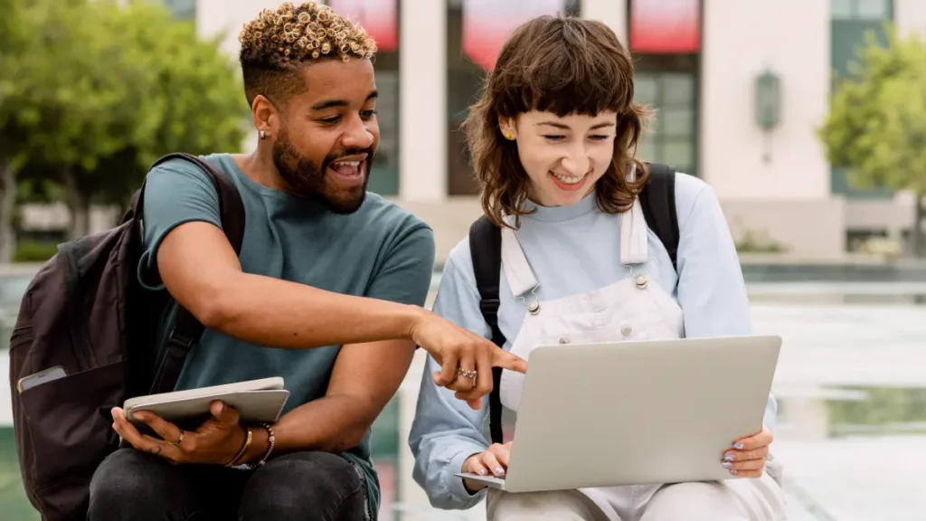 Jovens interagindo animados com notebook em área externa universitária.