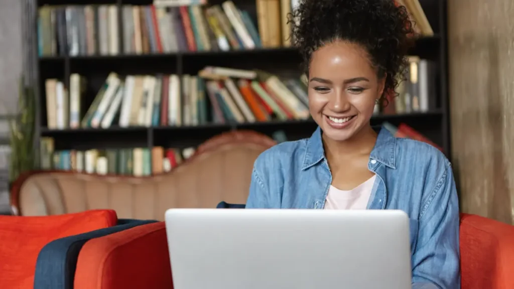 Jovem sorrindo usando notebook em sala com estante de livros.