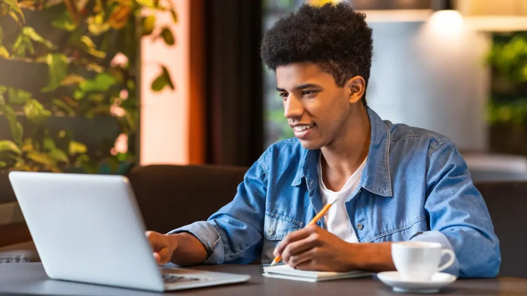 Jovem estudando em cafeteria com notebook e caderno aberto.