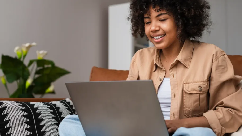 Jovem sorridente com cabelo cacheado usa notebook sentada em sofá com planta ao fundo.