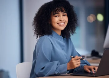Jovem profissional negra sorrindo enquanto trabalha em escritório moderno.