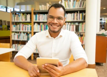 Jovem sorridente usando tablet em biblioteca moderna.