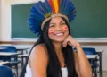 Mulher indígena com cocar de penas e pinturas vermelhas no rosto sentada em uma sala de aula, em referência à pós-graduação indígena.