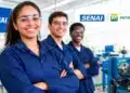Três jovens sorrindo em uma oficina industrial, com uniformes azuis e logotipos do SENAI e da Petrobras.