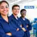 Três jovens sorrindo em uma oficina industrial, com uniformes azuis e logotipos do SENAI e da Petrobras.