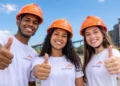 Três jovens usam camisetas da ArcelorMittal e capacetes laranjas enquanto fazem sinal de positivo para a câmera, com uma indústria ao fundo.
