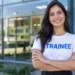 Jovem sorrindo, de braços cruzados, usando blusa branca com “TRAINEE” em azul, em frente a uma empresa.