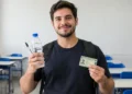 Jovem segura água, caneta e documento de identificação em sala de aula.