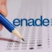 Mão preenche questão de múltipla escolha de prova do Enade com lápis azul ao lado do logo do exame.