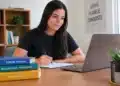 Jovem estuda com notebook, caderno e livros sobre formação profissional em ambiente iluminado.
