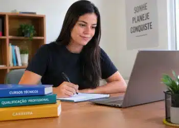 Jovem estuda com notebook, caderno e livros sobre formação profissional em ambiente iluminado.