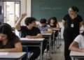 Alunos realizando o Enem 2026 com professora ao lado e um jovem levantando a mão, simbolizando as regras nos dias das provas do Enem 2026.