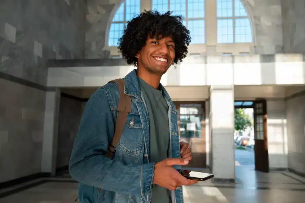 Jovem sorridente em hall de entrada iluminado segura celular.