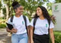 Duas estudantes sorrindo em um campus arborizado, com camisetas brancas do IFRJ, mochilas e livros.