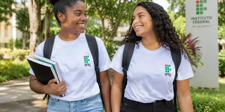 Duas estudantes sorrindo em um campus arborizado, com camisetas brancas do IFRJ, mochilas e livros.