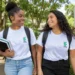 Duas estudantes sorrindo em um campus arborizado, com camisetas brancas do IFRJ, mochilas e livros.
