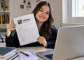 Jovem segura uma folha da Udesc em frente ao notebook, em um quarto claro e organizado.