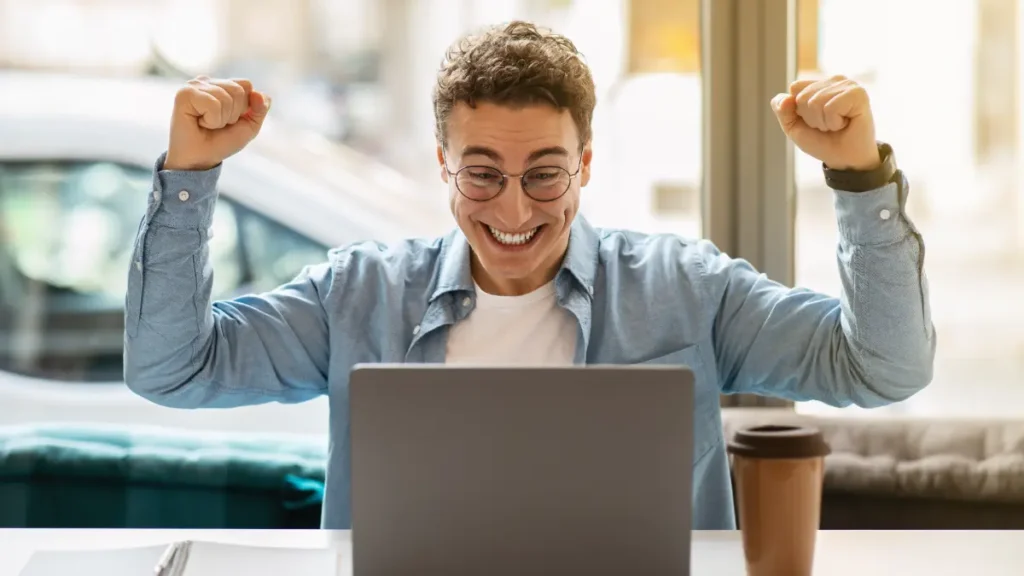 Homem comemora vitória usando notebook em ambiente moderno.