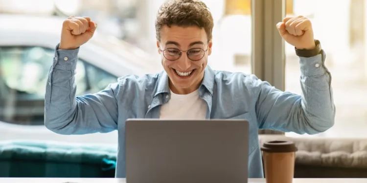 Homem comemora vitória usando notebook em ambiente moderno.