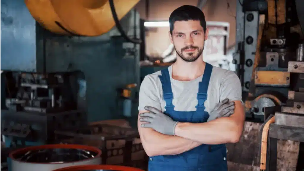 Serralheiro posando confiante em oficina industrial com maquinário.