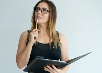 Mulher pensativa com óculos segura caderno e caneta.